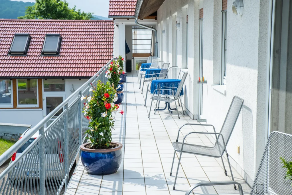 Sonniger Balkon mit Stühlen, gedeckten Tischen und blühenden Kübelpflanzen