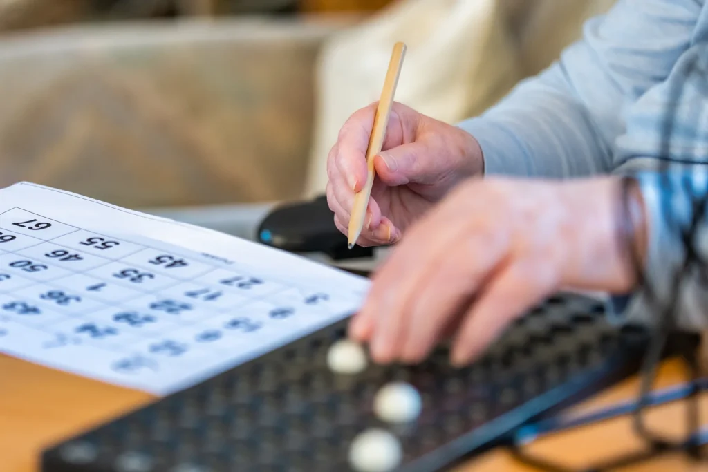 Hände beim Bingo am Tisch: ein Bingoschein, ein Bleistift und ein Steckbrett mit Kugeln