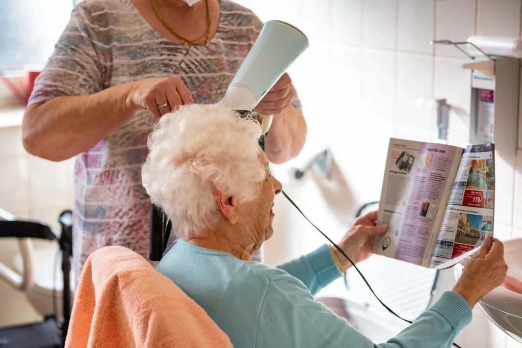 Eine Bewohnerin blättert in einer Zeitschrift, während ihr die Frisörin die weißen Haare föhnt