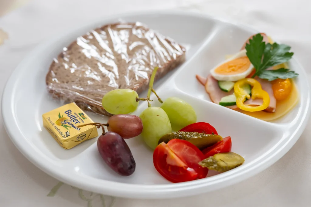 Teller mit Brot, Butter, Trauben, Tomaten, Gewürzgurke und einem Putenbruströllchen mit Ei