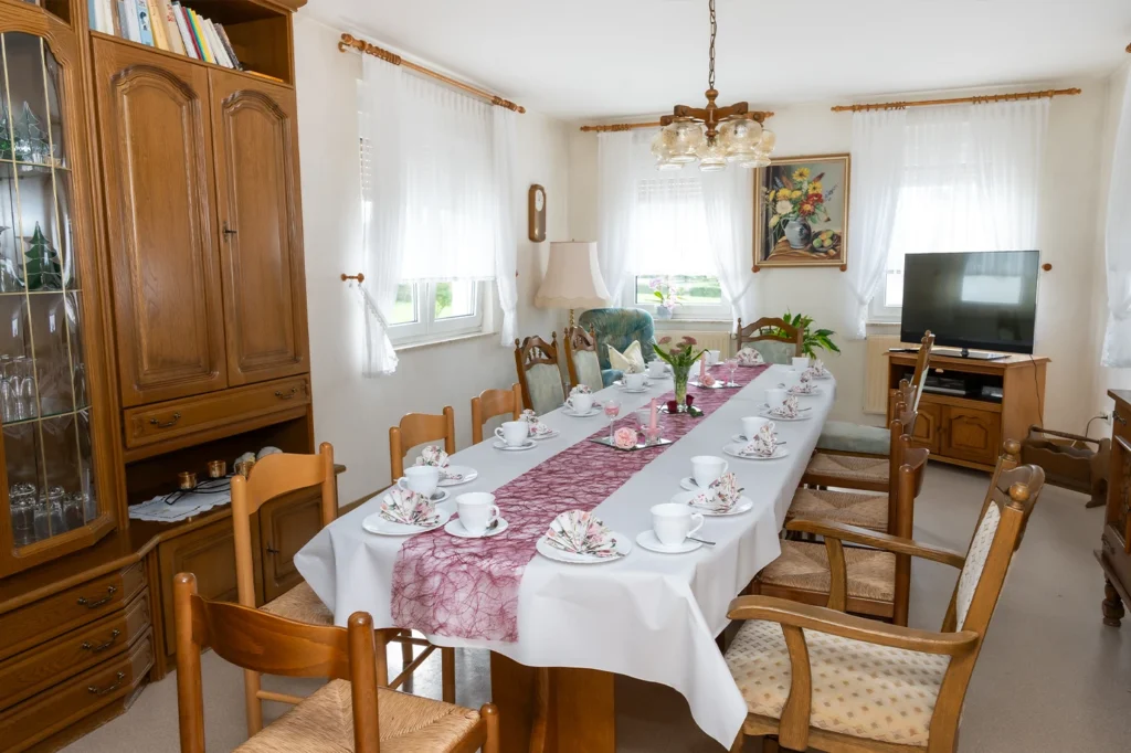 Wohnstube mit gedeckter Kaffeetafel, Holzstühlen, Wohnzimmerschrank und Fernseher