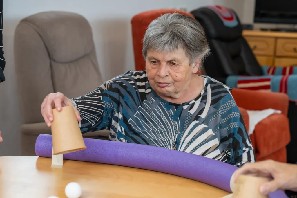 Eine Bewohnerin mit grauen Haaren setzt am Tisch einen Pappbecher auf eine lila Schwimmnudel