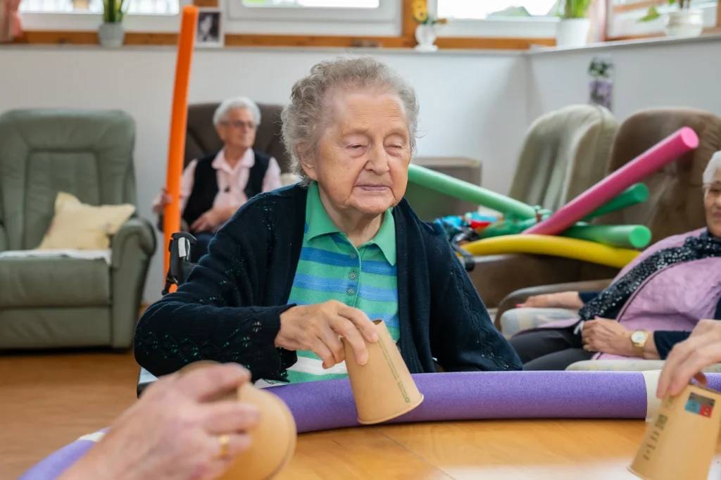 Eine Bewohnerin in dunkelblauer Strickjacke schiebt am Tisch einen Pappbecher an einer lila Schwimmnudel entlang