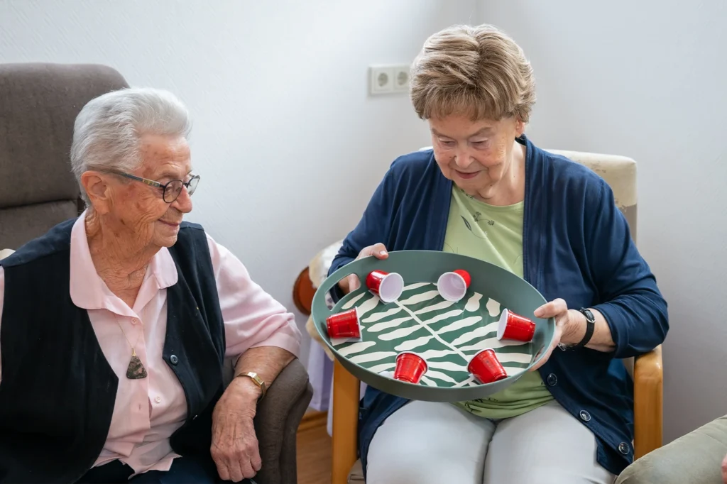 Zwei Bewohnerinnen sitzen nebeneinander, eine hält ein Tablett mit roten und weißen Bechern