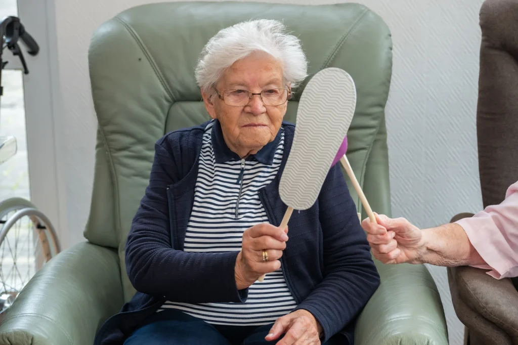 Eine Bewohnerin mit Brille hält im Sessel einen leichten Schläger hoch, eine zweite Hand reicht ihr einen weiteren