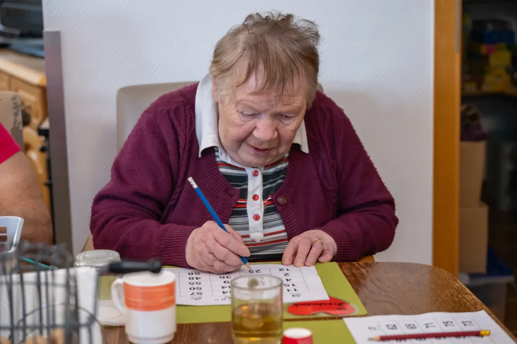 Eine Bewohnerin in weinroter Strickjacke löst am Tisch mit einem Bleistift eine Zahlenaufgabe