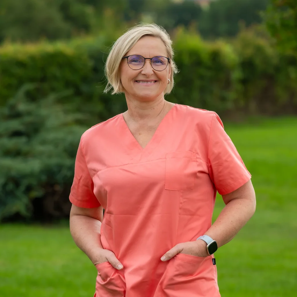 Mitarbeiterin mit kurzen blonden Haaren und Brille in lachsfarbenem Kasack