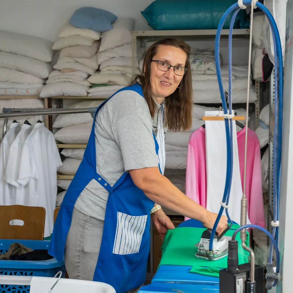 Mitarbeiterin in blauer Schürze bügelt in der Wäscherei, im Regal liegt gefaltete Wäsche