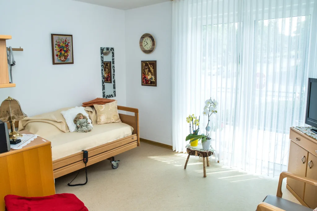 Bewohnerzimmer mit Pflegebett, eigenen Bildern an der Wand und bodentiefem Fenster zum Balkon